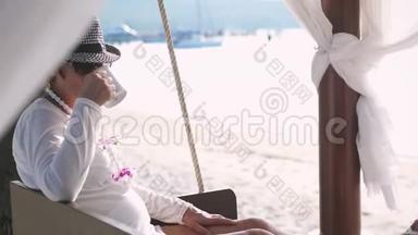 在美丽的海滩和秋千上，体贴的老妇人在休息时喝咖啡。 慢动作。 3840x2160
